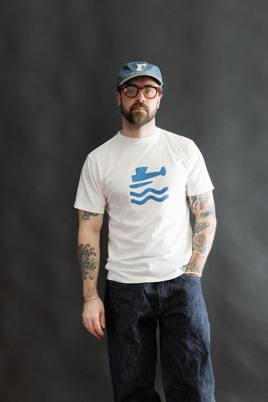 5.4oz Raffy Tenjiku Printed T-Shirt Seaplane - Off White worn by a man with tortoiseshell glasses, denim cap, showcasing vintage-inspired style with a seaplane graphic and tubular knit design.