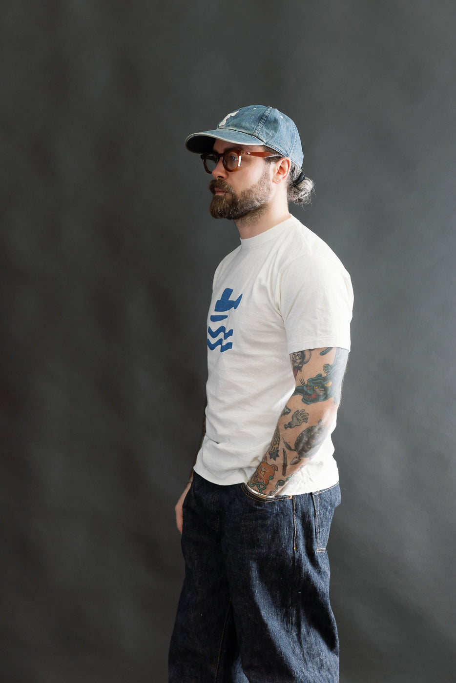 A bearded man models the 5.4oz Raffy Tenjiku Printed T-Shirt Seaplane in Off White, showcasing its American vintage-inspired design with a blue graphic and tubular construction, paired with dark denim jeans.