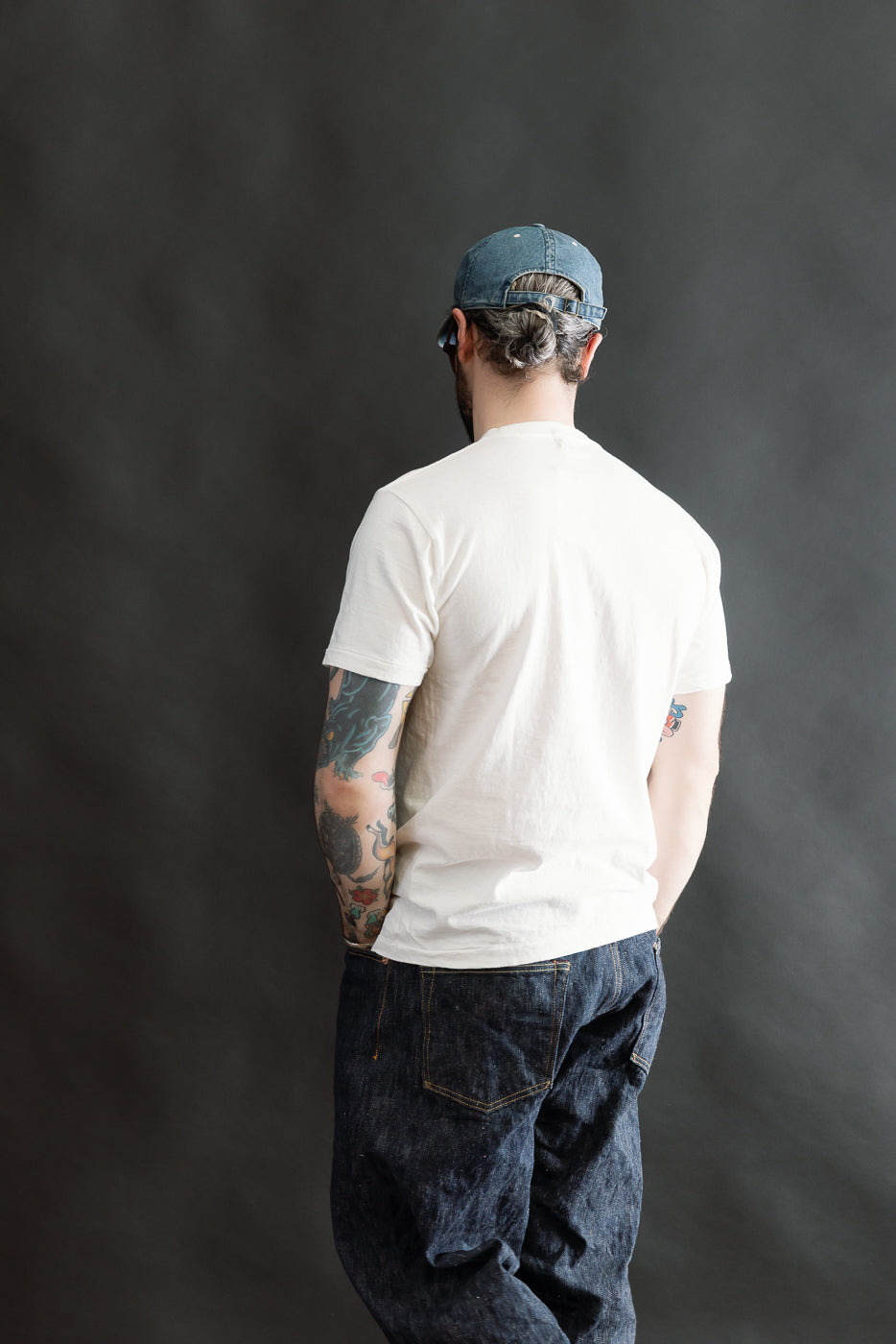 A male model showcases the 5.4oz Raffy Tenjiku Printed T-Shirt Seaplane in Off White, highlighting its vintage-inspired design, tubular construction, and unique slub texture, paired with jeans and a cap.