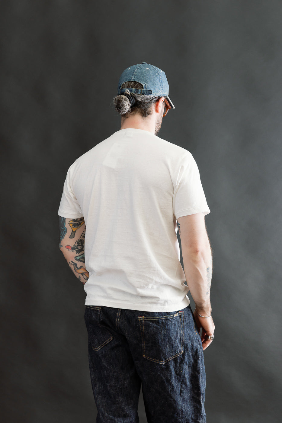 Back view of a man in a 5.4oz Raffy Tenjiku Printed T-Shirt Seaplane - Off White, highlighting its seamless tubular knit and vintage-inspired slub jersey texture, paired with denim jeans and cap.