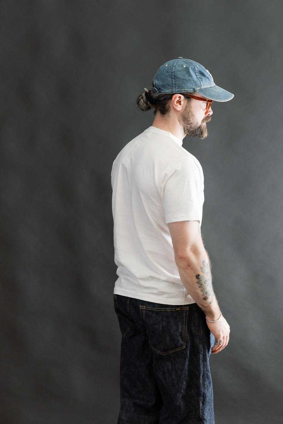 Man in profile wears the 5.4oz Raffy Tenjiku Printed T-Shirt Seaplane in off-white, styled with dark indigo jeans and a faded blue baseball cap, exemplifying premium vintage-inspired menswear.
