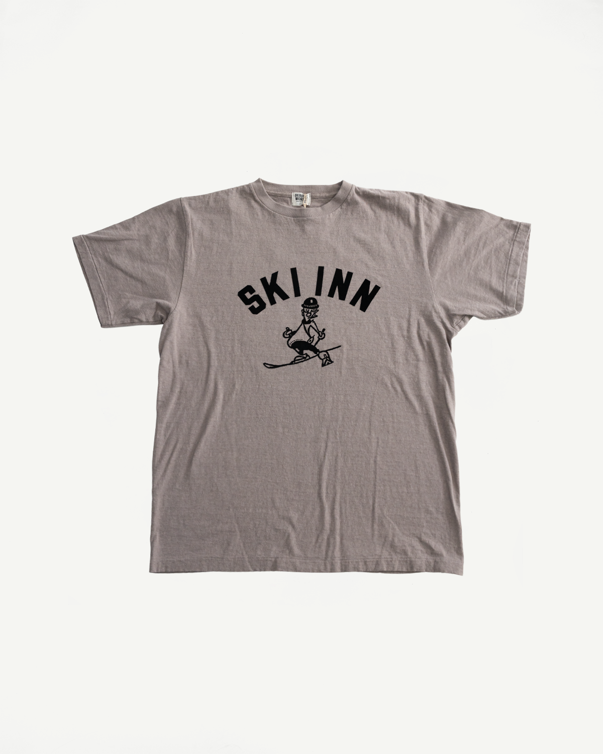 5.4oz - Raffy Tenjiku Printed T-Shirt Ski Inn - Grey features bold black text and a cartoon skier graphic on a gray crew-neck, highlighting vintage-inspired design with premium slub jersey fabric.