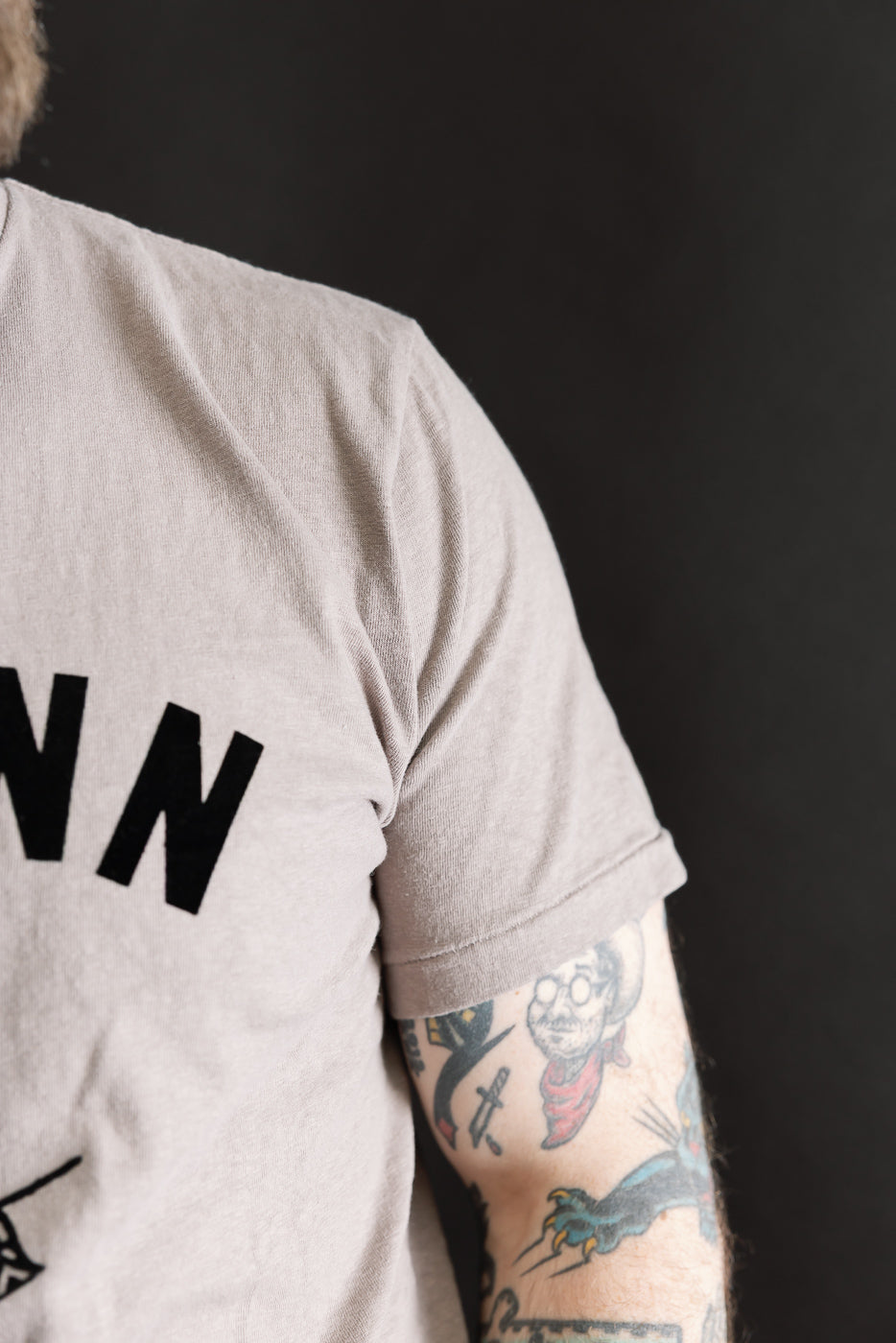 The 5.4oz Raffy Tenjiku Printed T-Shirt Ski Inn in Grey features bold black lettering on the visible half. A tattooed forearm with vibrant designs adds a unique touch to the presentation.