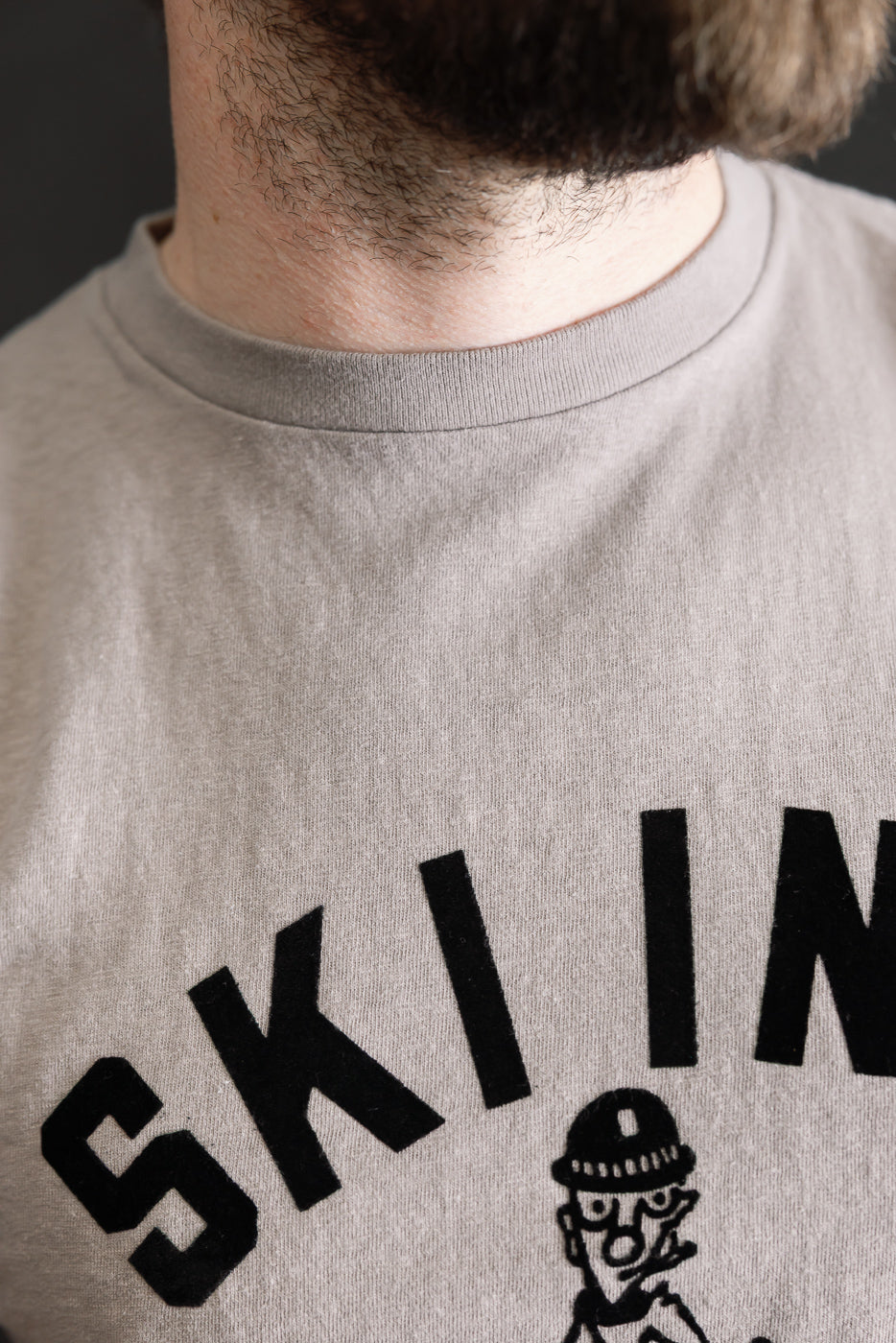 Close-up of a man's lower face and upper torso wearing the 5.4oz Raffy Tenjiku Printed T-Shirt Ski Inn in grey, showcasing bold black lettering and a cartoon character on its textured organic cotton fabric.