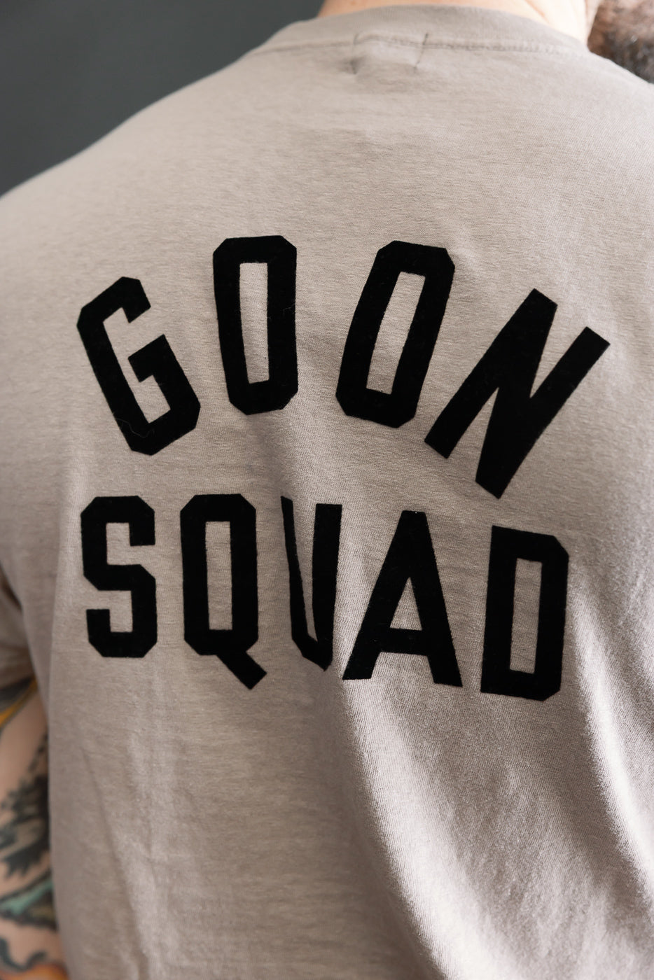Close-up of a light gray 5.4oz Raffy Tenjiku Printed T-Shirt Ski Inn featuring bold black GOON SQUAD text, showcasing the slub jersey texture and part of a tattooed forearm.