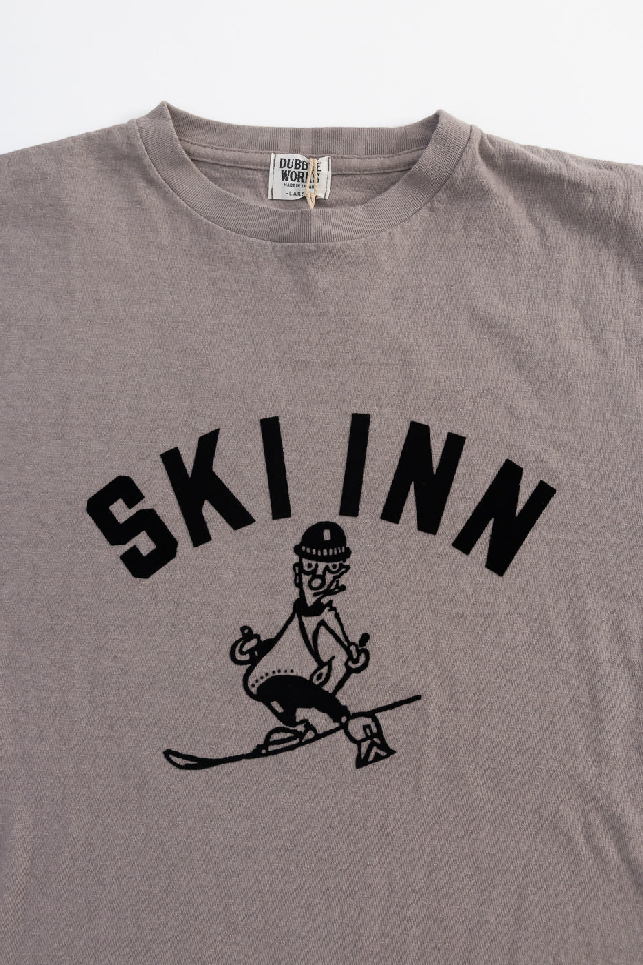 5.4oz - Raffy Tenjiku Printed T-Shirt Ski Inn - Grey, featuring a cartoon skier and SKI INN text, crafted in Japan using organic cotton with a nep-like texture and seamless tubular construction.