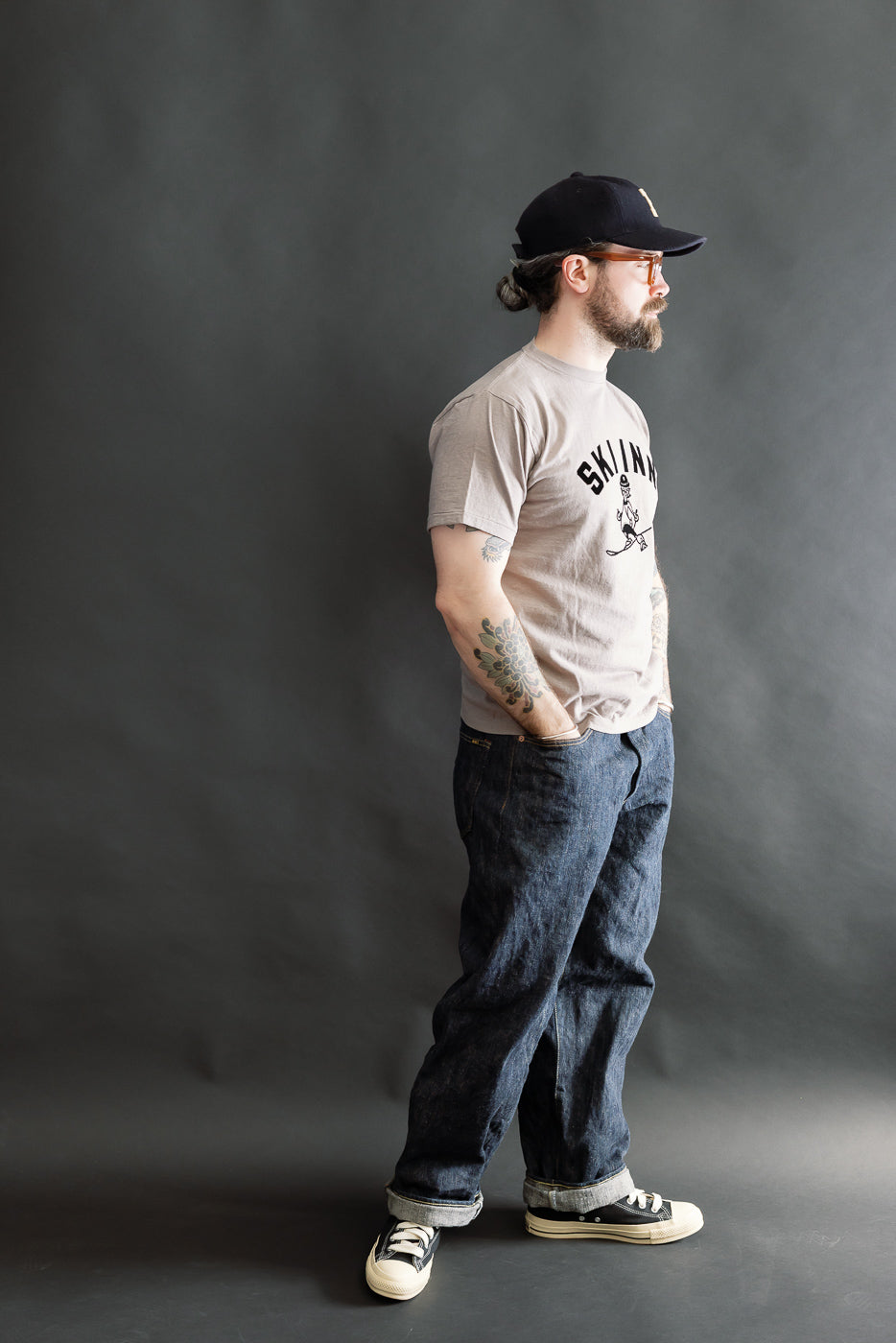 The 5.4oz Raffy Tenjiku Printed T-Shirt Ski Inn in grey is worn by a bearded man, showcasing its vintage-inspired design and nep-like texture, complemented by dark denim jeans and sneakers.