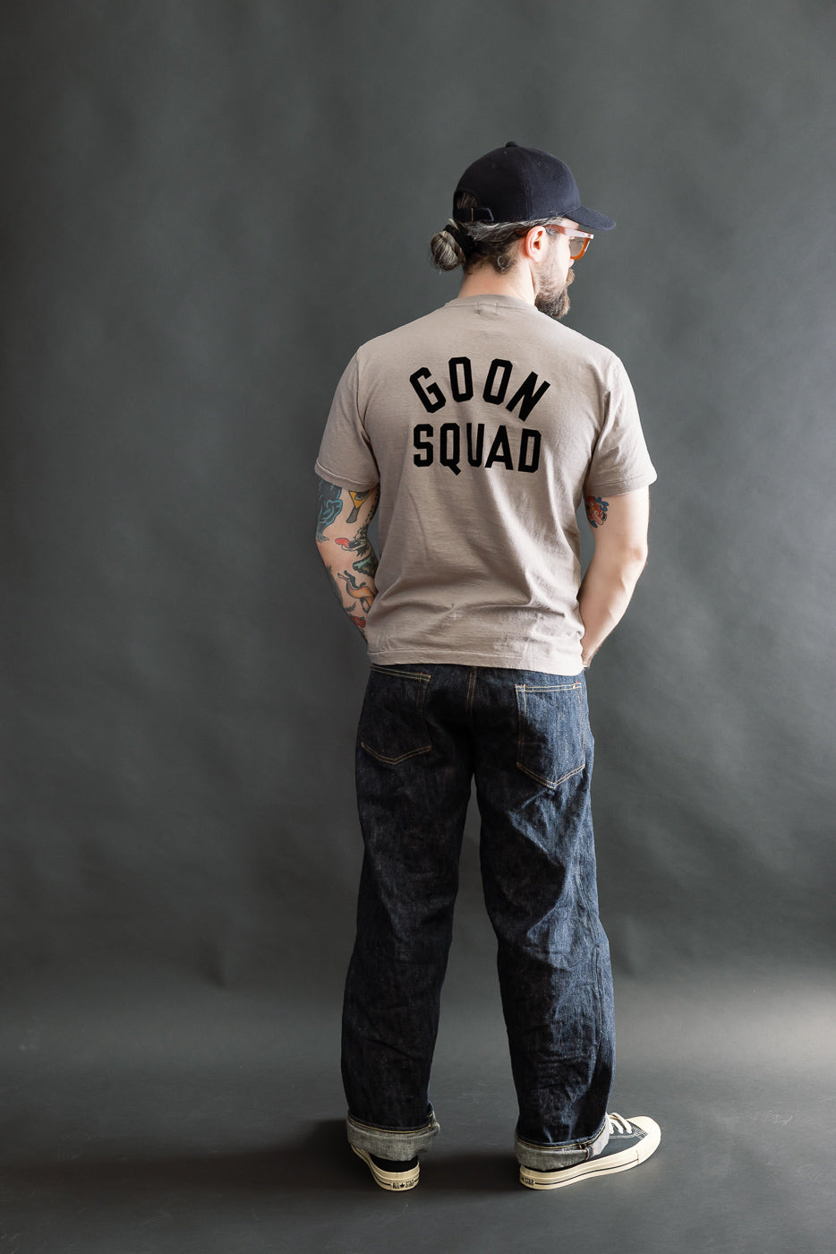 Man wearing 5.4oz Raffy Tenjiku Printed T-Shirt Ski Inn - Grey, showcasing GOON SQUAD text. Paired with cuffed denim jeans and canvas sneakers, embodying Dant's premium menswear style.