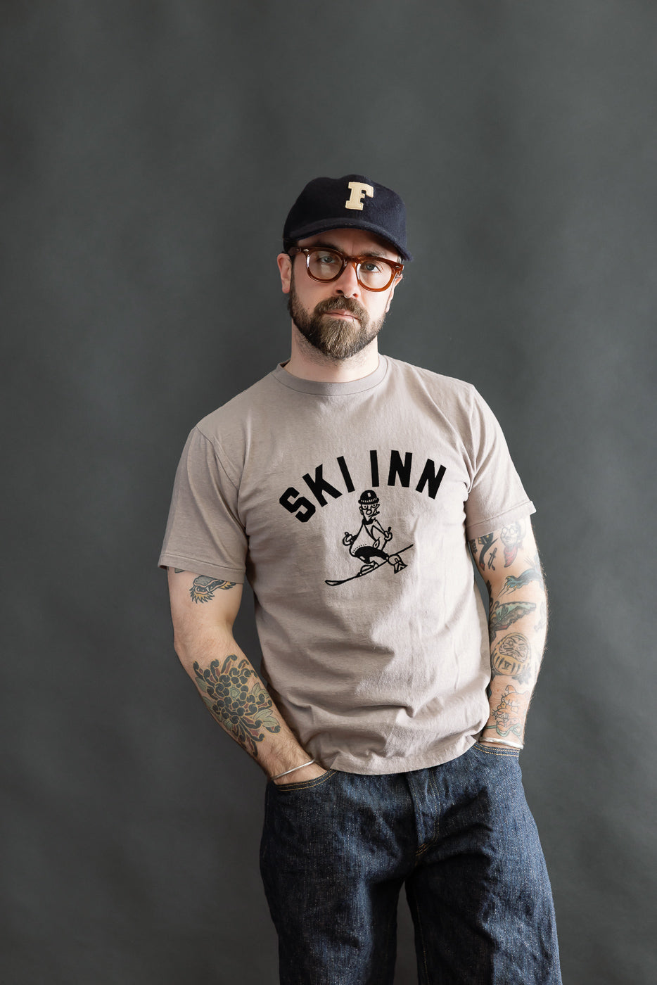 5.4oz Raffy Tenjiku Printed T-Shirt Ski Inn - Grey worn by a casually dressed man, highlighting its vintage-inspired design, organic slub jersey texture, and seamless tubular construction for a classic, relaxed fit.
