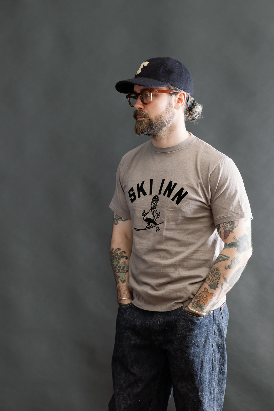 Man wears the 5.4oz Raffy Tenjiku Printed T-Shirt Ski Inn – Grey, showcasing its vintage-inspired design and slim fit. The light brown tee features a cartoon skier graphic, paired with dark denim jeans.