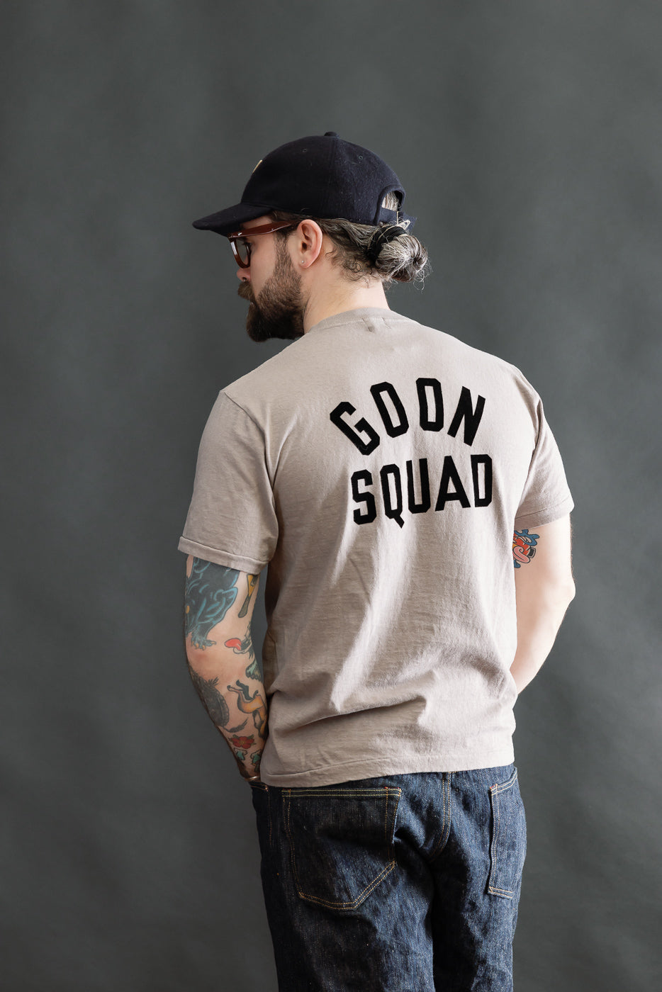 The 5.4oz Raffy Tenjiku Printed T-Shirt Ski Inn - Grey features bold GOON SQUAD lettering on the back, worn by a bearded man with tattoos, embodying vintage American style.