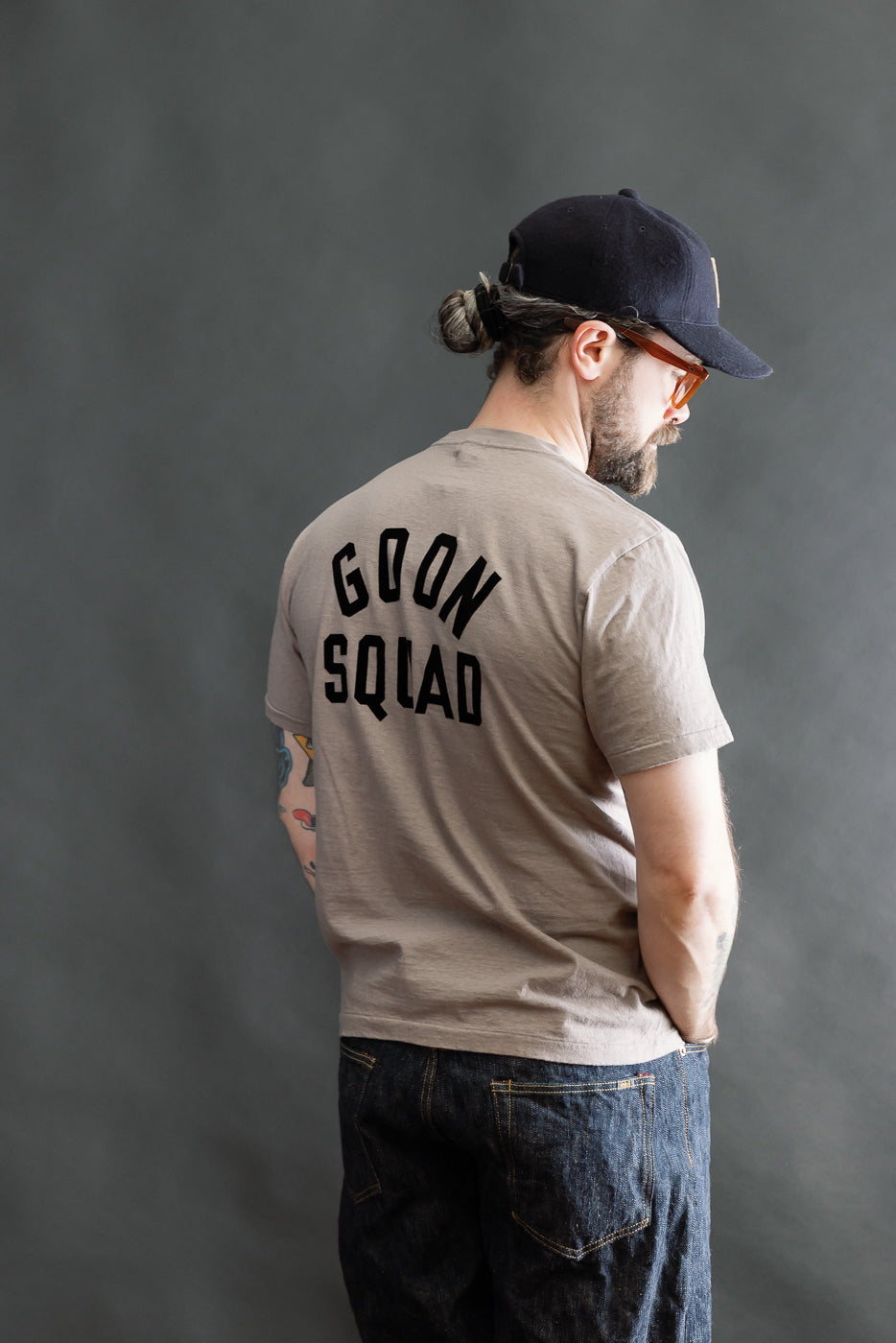 Man models the 5.4oz Raffy Tenjiku Printed T-Shirt Ski Inn - Grey by DUBBLE WORKS, showcasing its back design, tubular knit, and vintage-inspired slub jersey fabric with GOON SQUAD text.