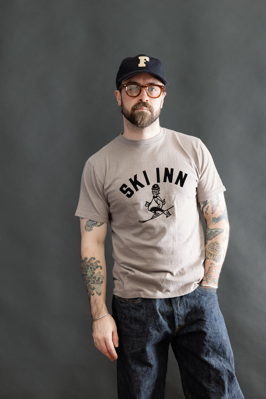 Man wearing the 5.4oz Raffy Tenjiku Printed T-Shirt Ski Inn - Grey, showcasing its vintage-inspired design, round neckline, and tubular construction. His style complements the heritage fashion ethos of Dant.