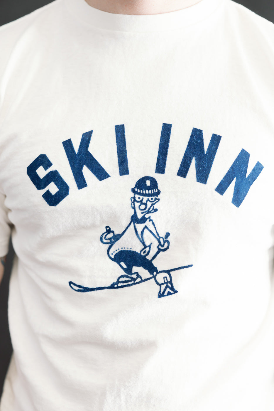 Close-up of the 5.4oz Raffy Tenjiku Printed T-Shirt Ski Inn - Off White, featuring SKI INN in bold blue block letters and a line drawing of a skier on high-quality, textured fabric.