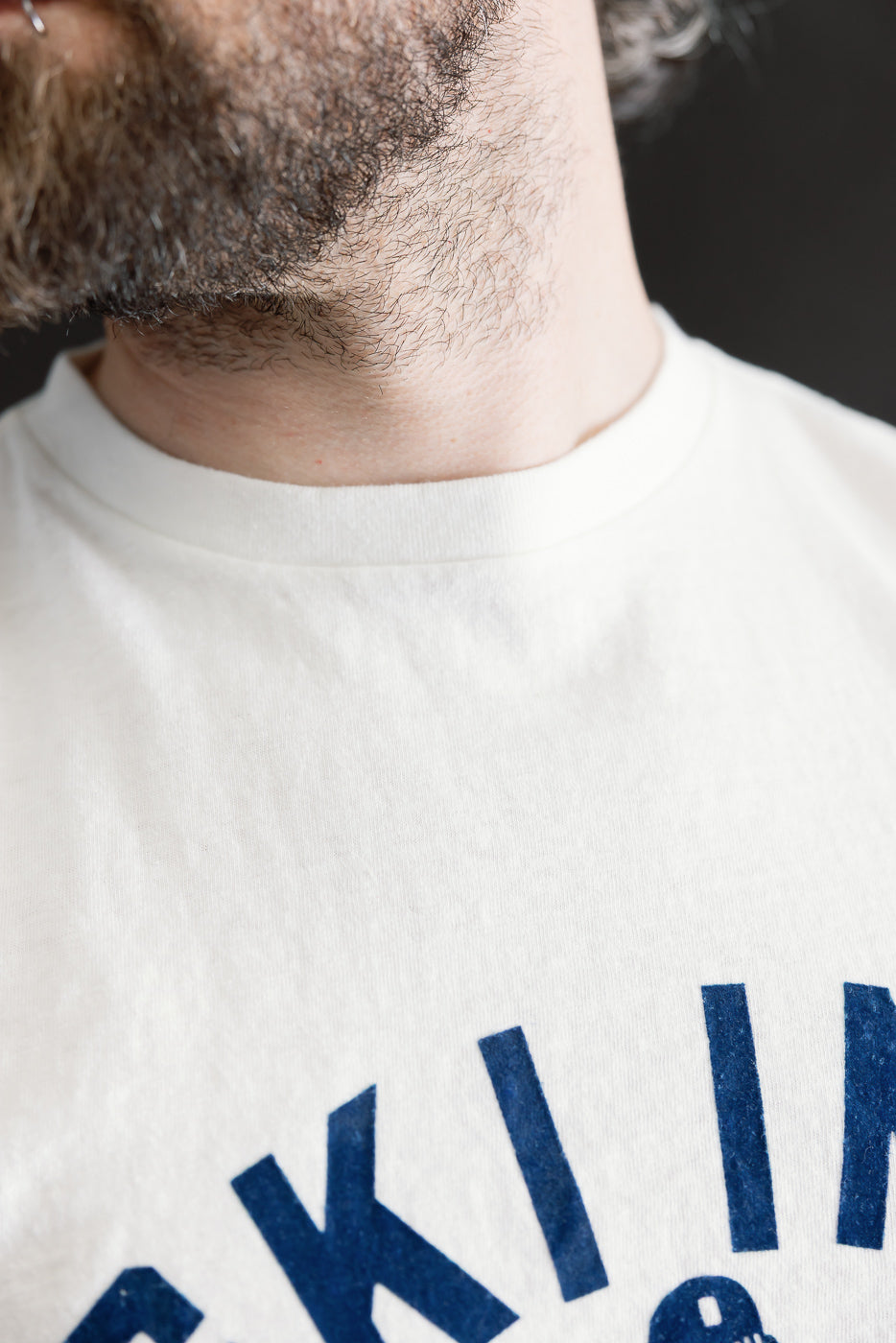 Close-up of a bearded man's lower face wearing the 5.4oz Raffy Tenjiku Printed T-Shirt Ski Inn - Off White, highlighting the vintage-inspired, nep-textured fabric with visible blue graphic.