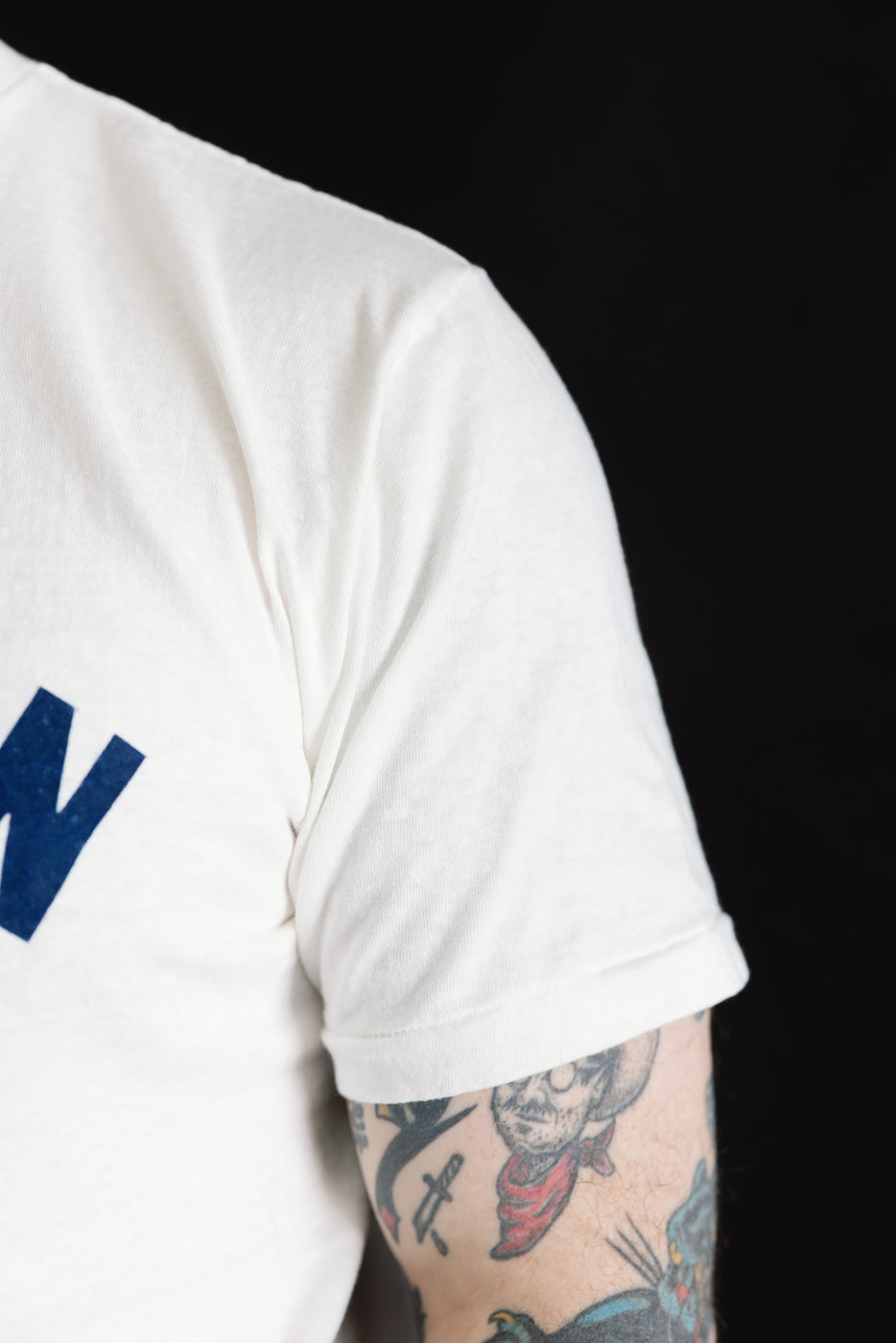 The 5.4oz Raffy Tenjiku Printed T-Shirt Ski Inn features a vintage-inspired design with a stylized cowboy tattoo, highlighting its seamless, tubular construction and organic cotton blend for a unique texture.