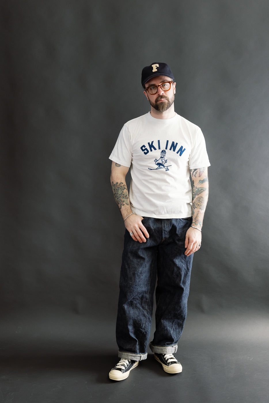 Man wearing the 5.4oz Raffy Tenjiku Printed T-Shirt Ski Inn in Off White, styled with loose dark denim jeans and canvas sneakers, showcasing visible forearm tattoos against a plain background.
