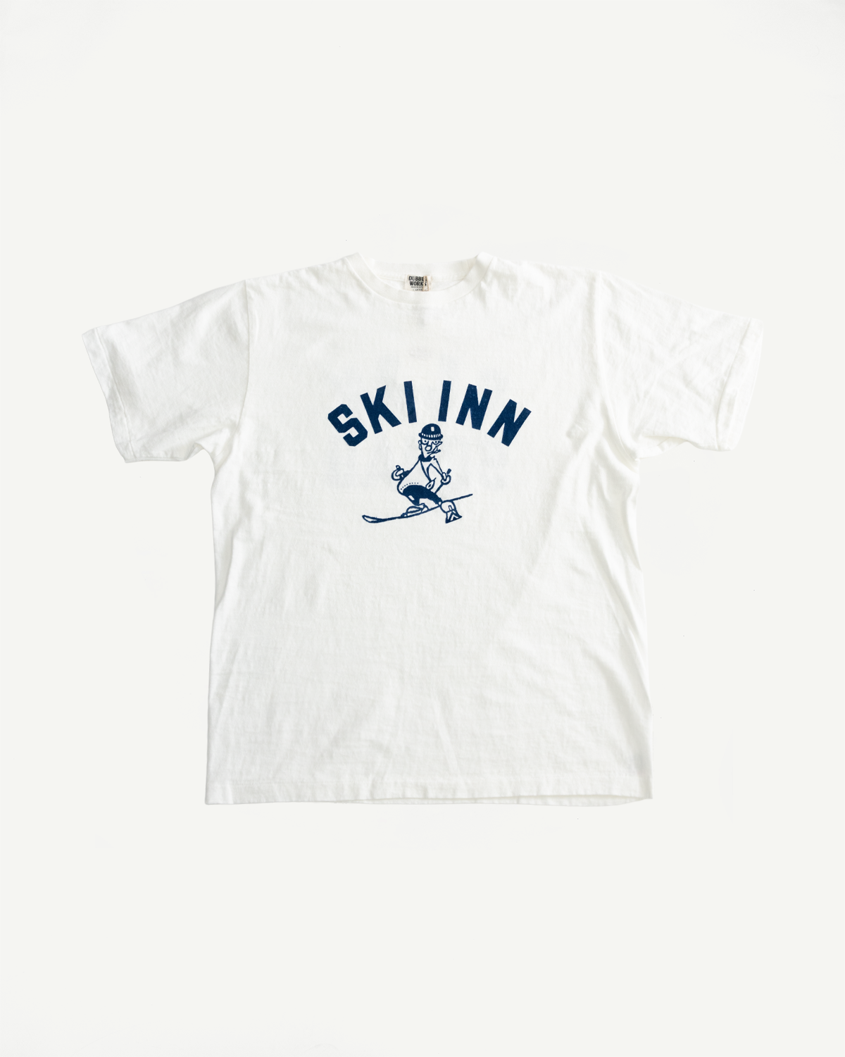 The 5.4oz Raffy Tenjiku Printed T-Shirt Ski Inn - Off White features a blue SKI INN graphic with a cartoon skier on a vintage-inspired white T-shirt.