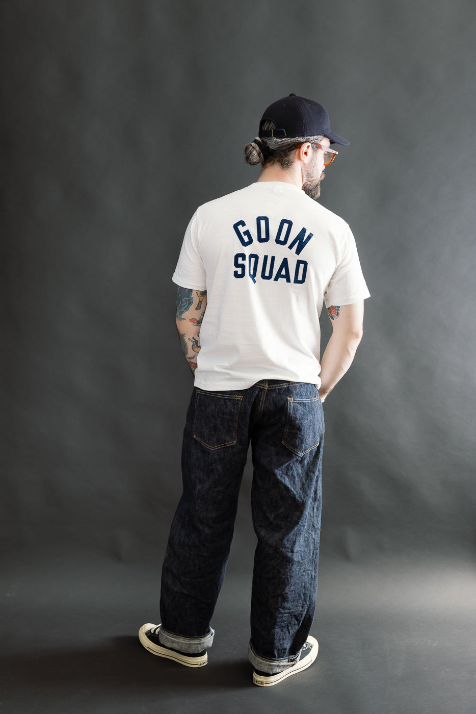 A man showcases the 5.4oz - Raffy Tenjiku Printed T-Shirt Ski Inn - Off White, featuring GOON SQUAD text, paired with denim jeans, sneakers, and a baseball cap.