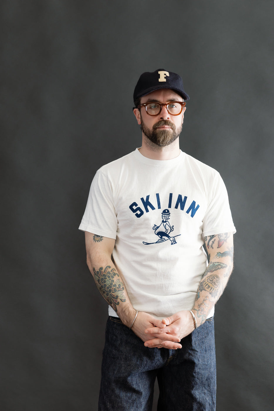 Bearded man in a 5.4oz Raffy Tenjiku Printed T-Shirt Ski Inn - Off White, featuring a vintage-inspired design with a skier graphic. Styled with dark jeans and a navy cap, showcasing a heritage look.