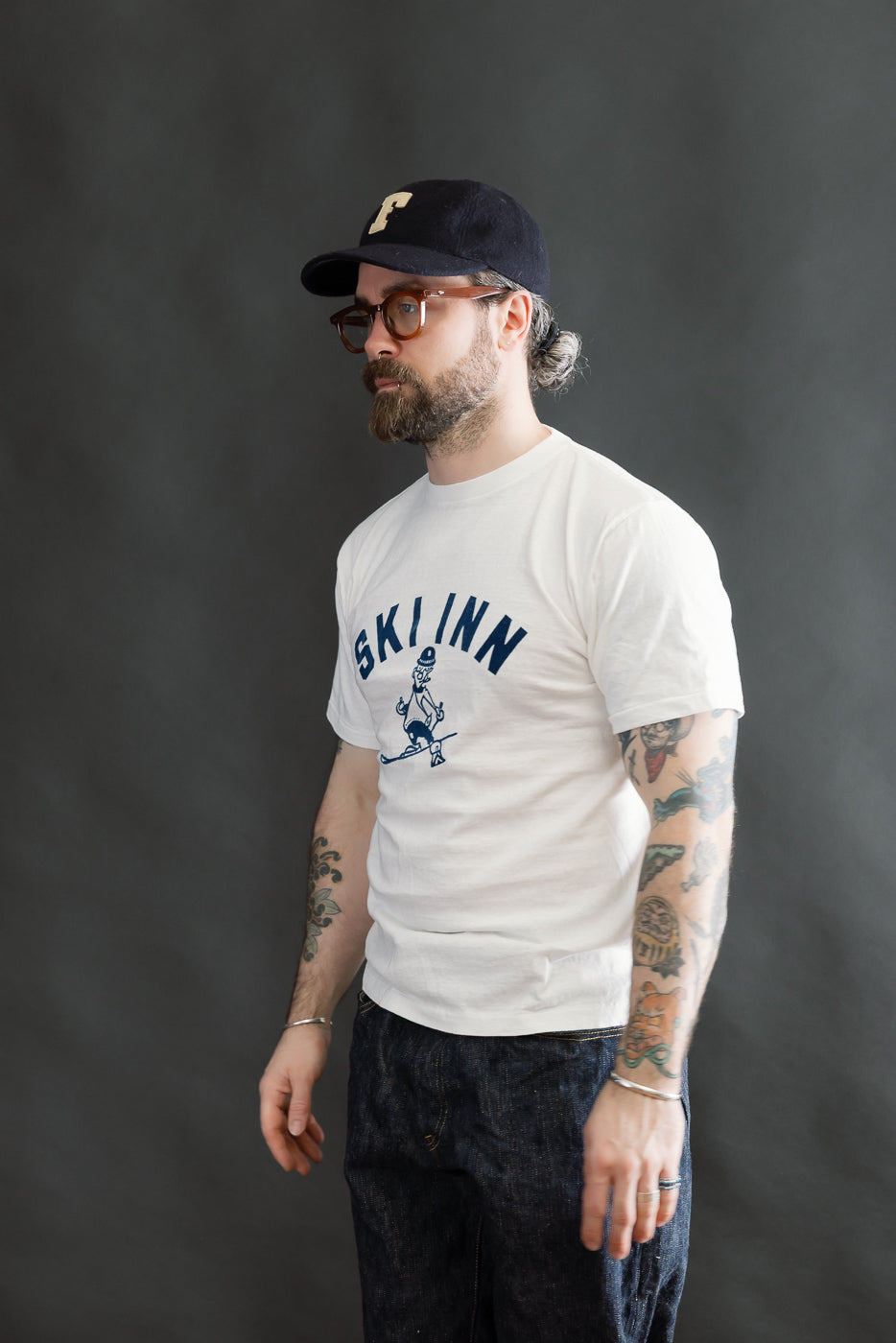 A man models the 5.4oz Raffy Tenjiku Printed T-Shirt Ski Inn - Off White, showcasing its vintage-style design, tubular knit, and slub jersey texture, paired with denim jeans and accessories.