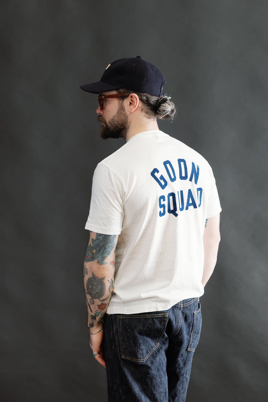 A bearded man models the 5.4oz Raffy Tenjiku Printed T-Shirt Ski Inn in Off White, showcasing its vintage-inspired style with GOON SQUAD in blue across the back.