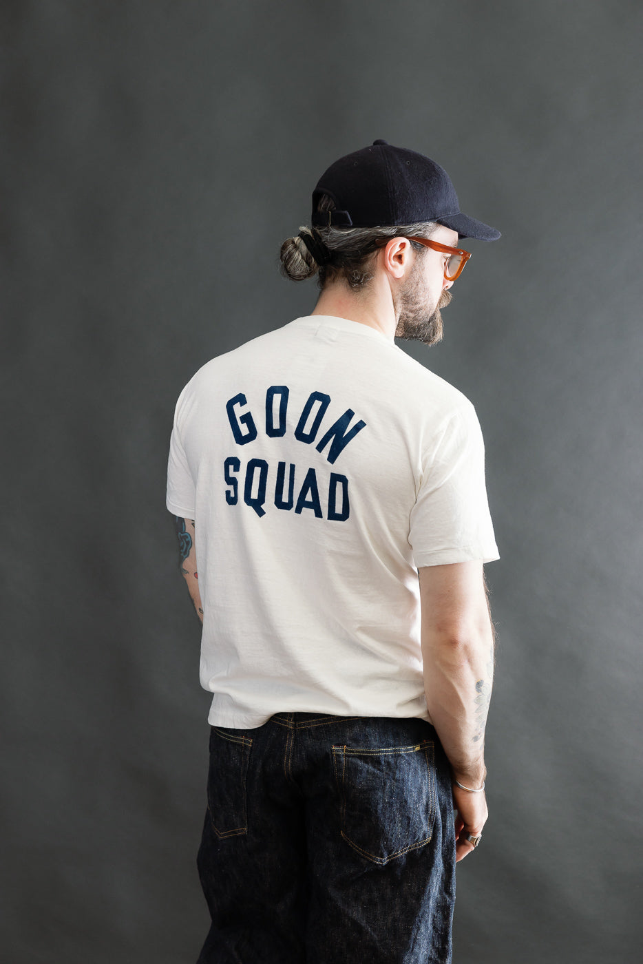 A man models the 5.4oz Raffy Tenjiku Printed T-Shirt Ski Inn in Off White, showcasing vintage-inspired design with GOON SQUAD print, paired with dark denim jeans and a black cap.