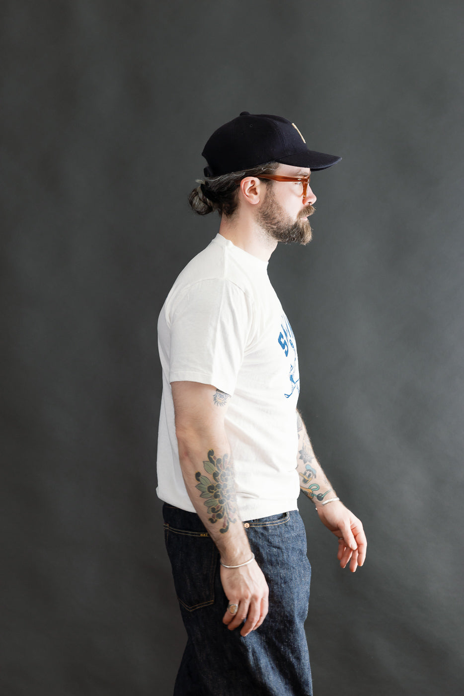 A bearded man wears a 5.4oz Raffy Tenjiku Printed T-Shirt Ski Inn - Off White, showcasing its tubular knit and flock print design. His casual pose highlights the tee's vintage-inspired style.