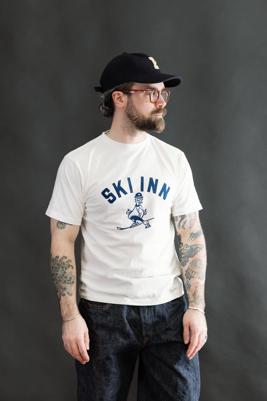 Man wearing 5.4oz Raffy Tenjiku Printed T-Shirt Ski Inn - Off White, showcasing vintage-inspired design with tubular knit, colorful tattoos visible, styled with clear glasses and baseball cap, embodying Dant's premium menswear style.