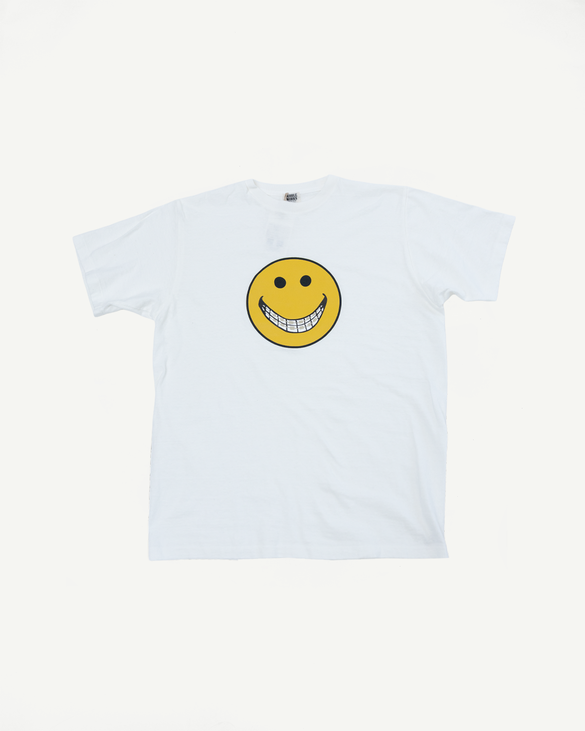 Flat lay of the front of a white t-shirt featuring a yellow smiley face with teeth.