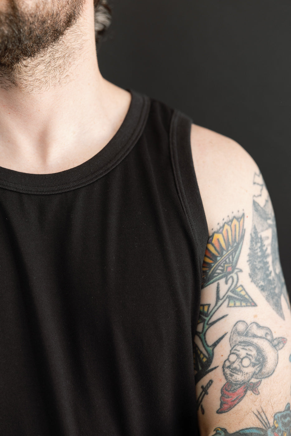 Close-up of male model's shoulder, showing black tank top with ribbed neckline and tattooed arm.