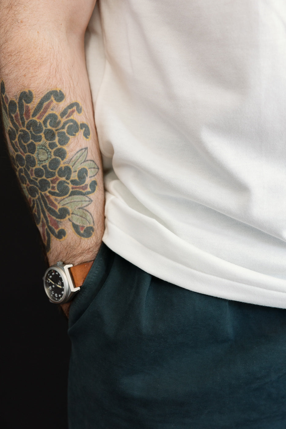 Close-up of male model's arm with floral tattoo, white tee, and dark pants, showcasing fabric texture and fit.