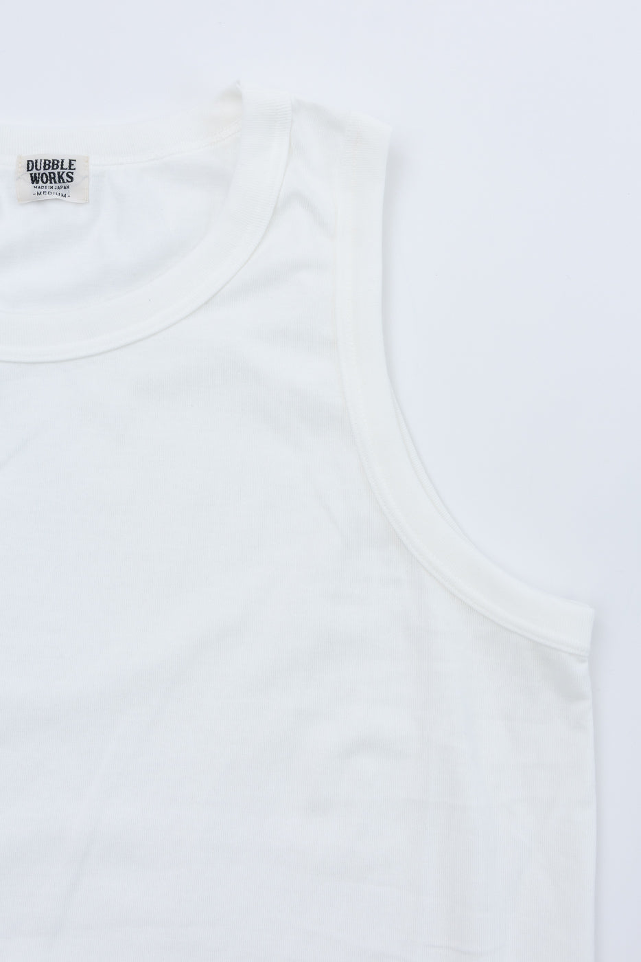 Close-up front view of white Dubble Works tank top showing tag, ribbed crew neck, and sleeve hem detail
