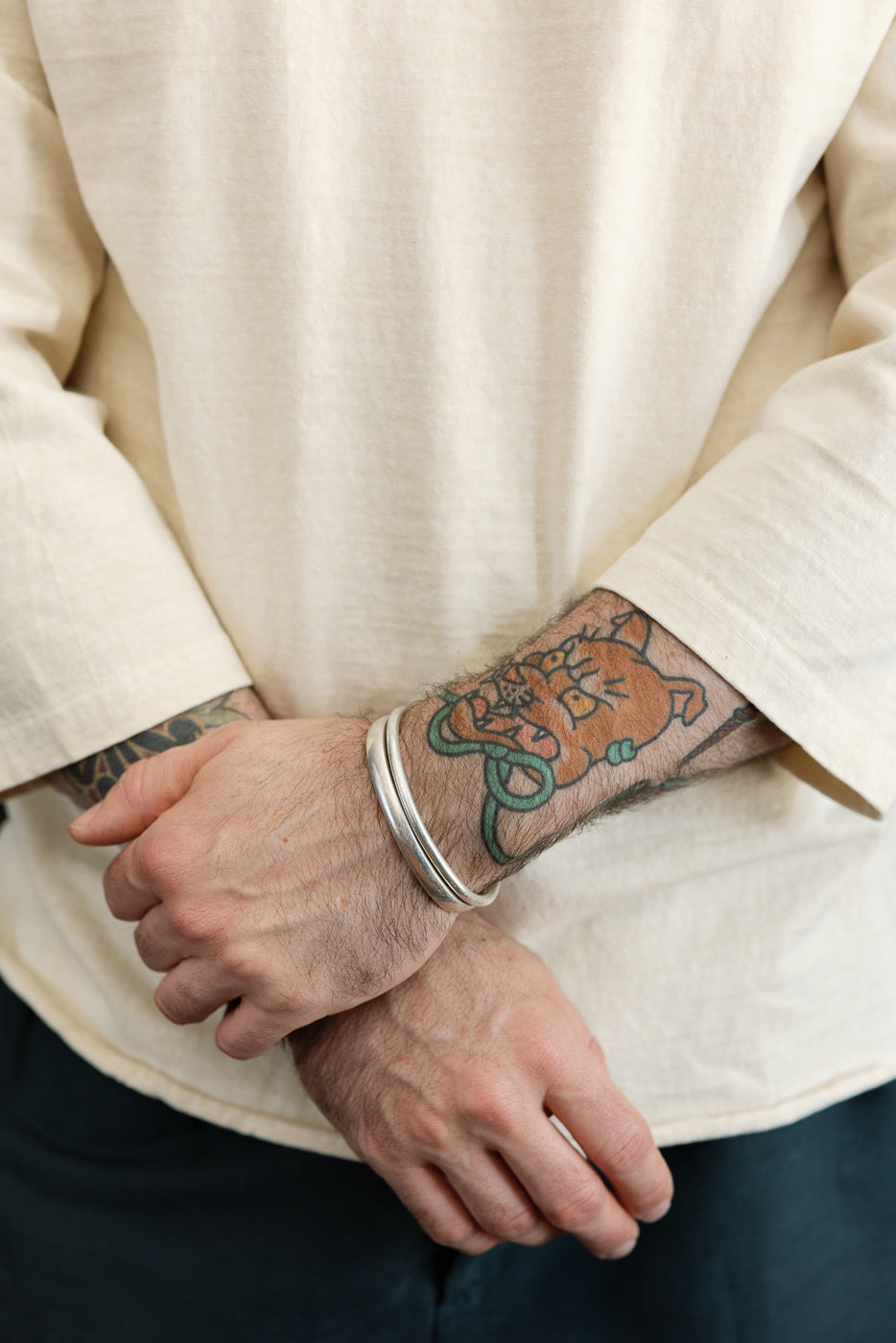 Close-up front view of male model's forearm showing tattoo, silver bracelet, and cream long-sleeve shirt cuff