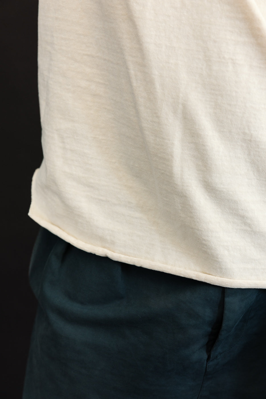 Close-up side view of male model's off-white tee hem, showing soft fabric texture and curved hemline over dark pants