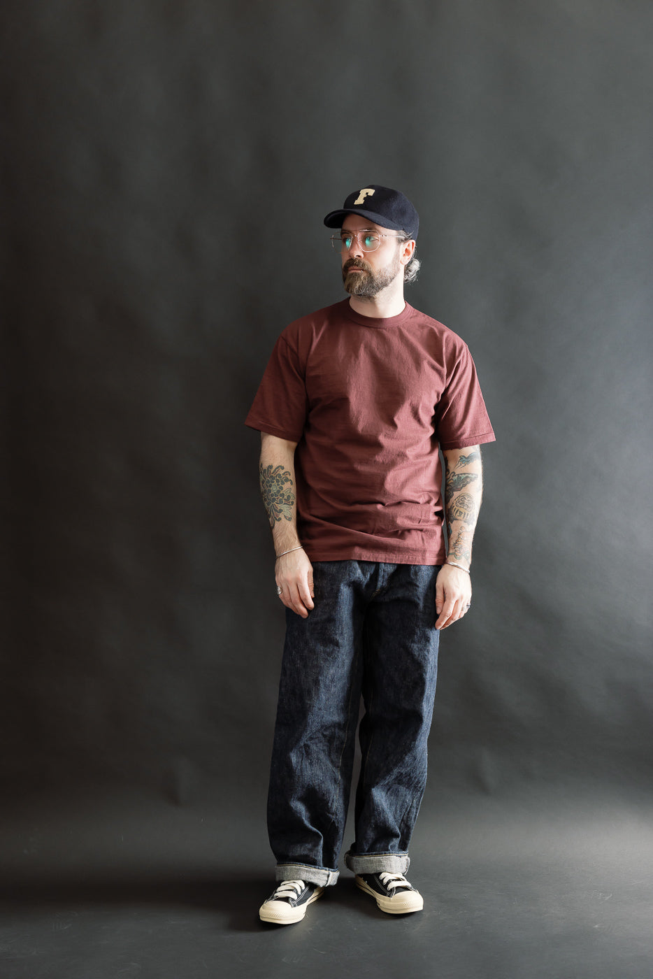 A casually dressed man models the 7.7oz Fallen Cotton Midweight T-Shirt S/S - Bordeaux, showcasing its short sleeves and soft, textured recycled cotton fabric, styled with dark denim and sneakers.