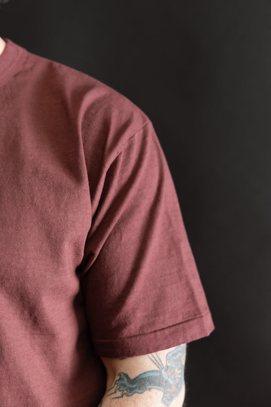 Close-up of the 7.7oz - 'Fallen Cotton' Midweight T-Shirt S/S - Bordeaux, featuring a maroon short-sleeve design with visible textured slub fabric and a tattooed forearm, embodying a casual yet refined look.