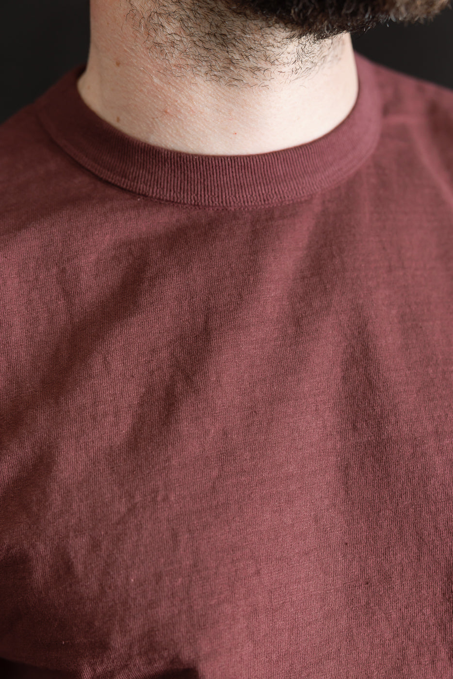 Close-up of a person wearing the 7.7oz Fallen Cotton Midweight T-Shirt S/S - Bordeaux, showcasing its maroon color, crew-neck design, and textured recycled cotton slub jersey fabric for a lived-in feel.