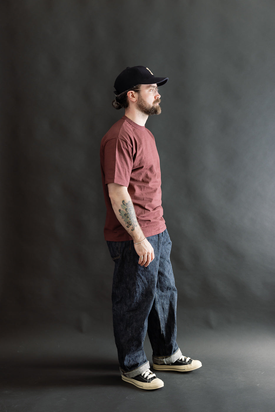 A bearded man models the 7.7oz Fallen Cotton Midweight T-Shirt S/S - Bordeaux, highlighting its classic fit and natural slub texture, styled with indigo jeans and sneakers.