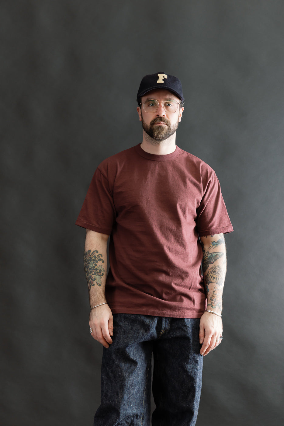 Man wearing the 7.7oz Fallen Cotton Midweight T-Shirt S/S - Bordeaux, showcasing its classic tee design with a round body and single-stitched hem, paired with jeans and a navy baseball cap.