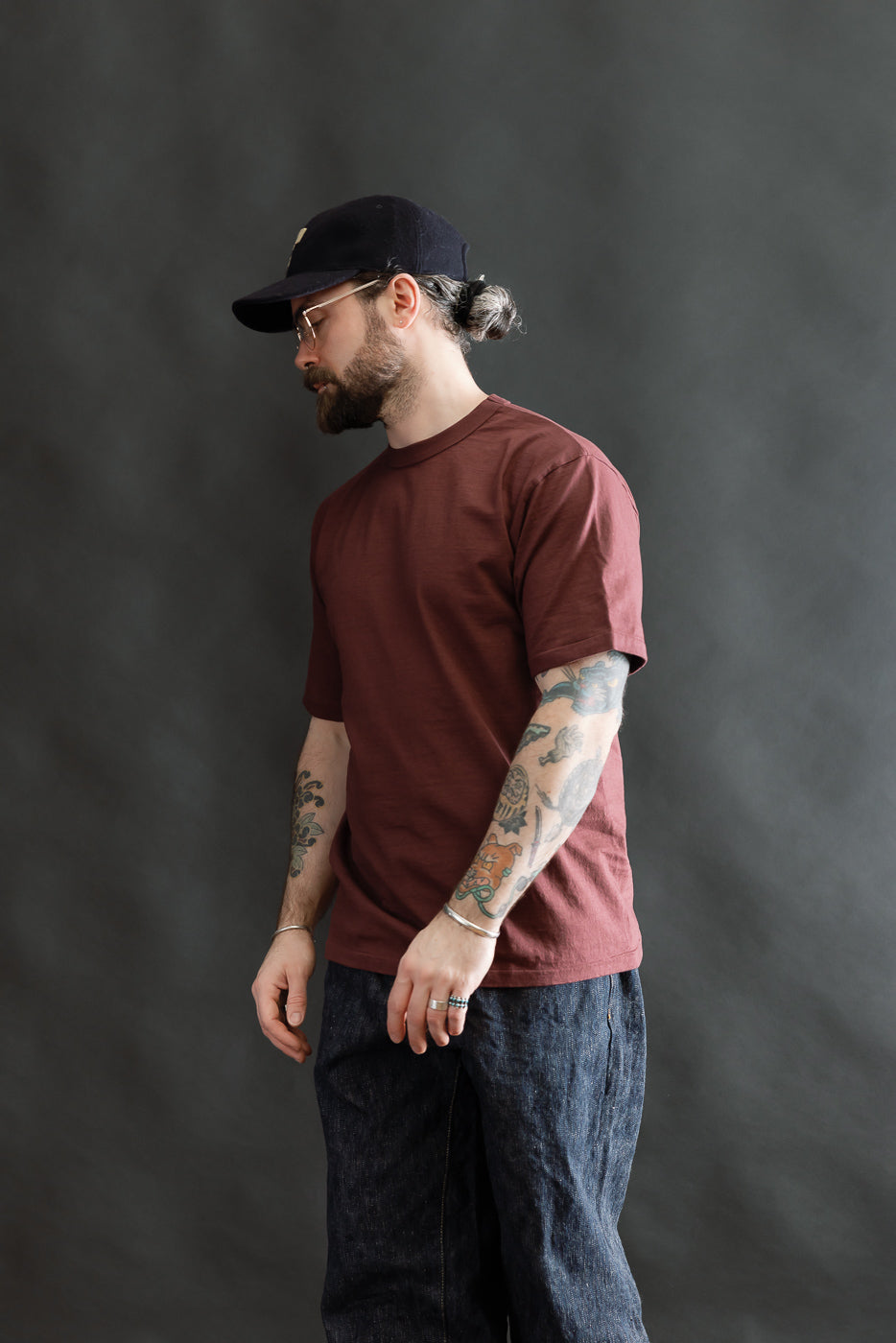 Man in a Fallen Cotton Midweight T-Shirt S/S - Bordeaux, featuring colorful tattoos, wearing loose-cut dark denim jeans, a dark baseball cap, and wire-frame glasses, with a contemplative look.
