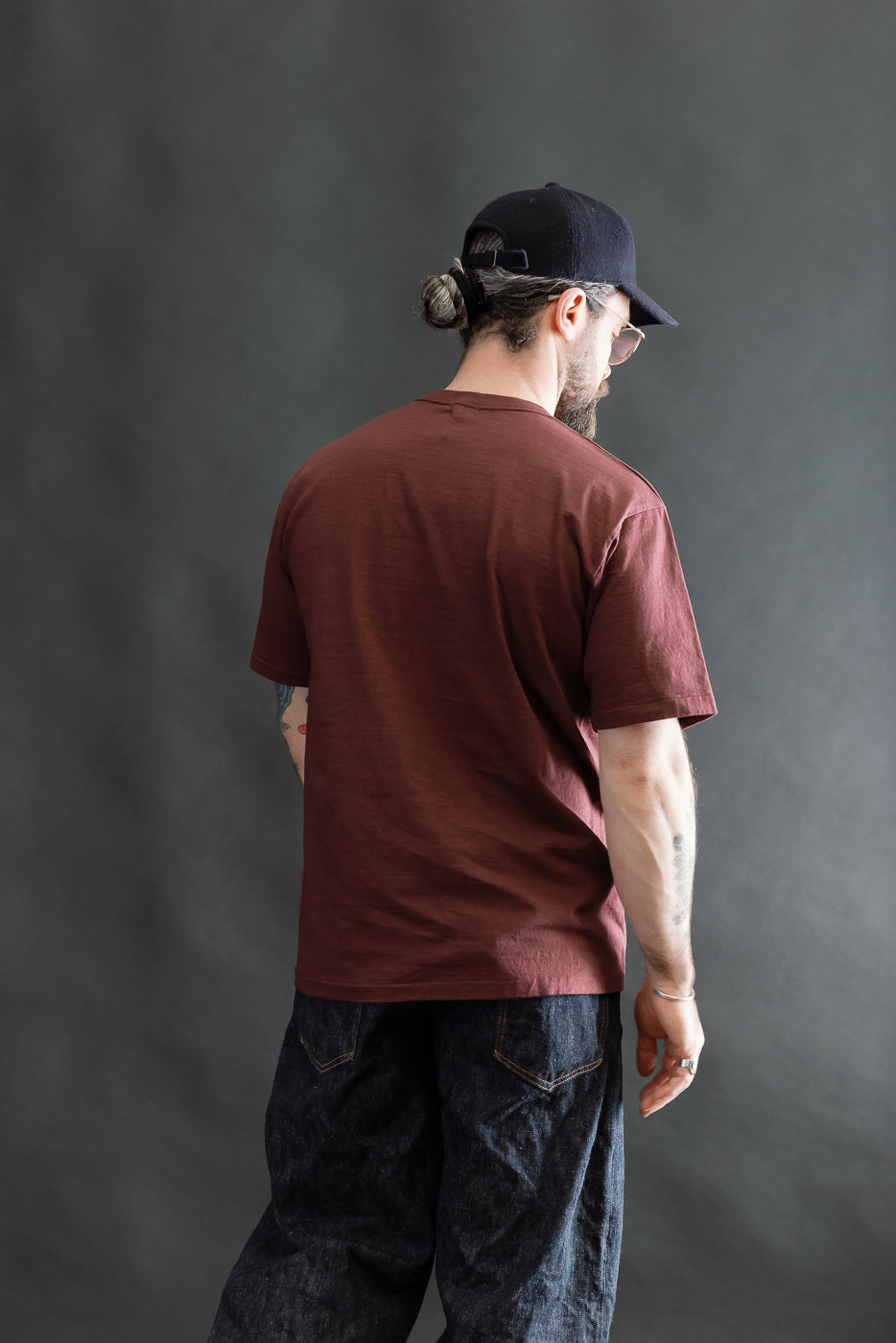 Man wearing a 7.7oz - Fallen Cotton Midweight T-Shirt S/S - Bordeaux, styled with loose dark blue denim jeans and a black backwards baseball cap, showcasing a blend of comfort and durability.