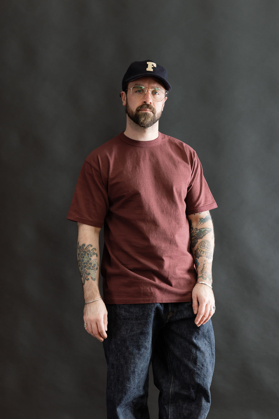 Model wearing the 7.7oz Fallen Cotton Midweight T-Shirt S/S - Bordeaux, showcasing its recycled cotton slub jersey's texture, relaxed fit, and classic tee construction, complete with tattoos and glasses.
