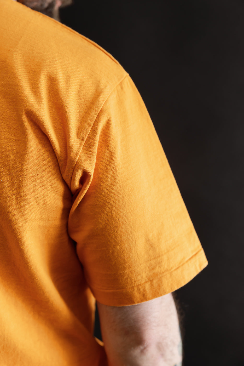 Close-up of a person wearing the 7.7oz - 'Fallen Cotton' Midweight T-Shirt S/S - Fade Orange, showcasing the textured recycled cotton slub jersey and classic tee construction against a dark backdrop.