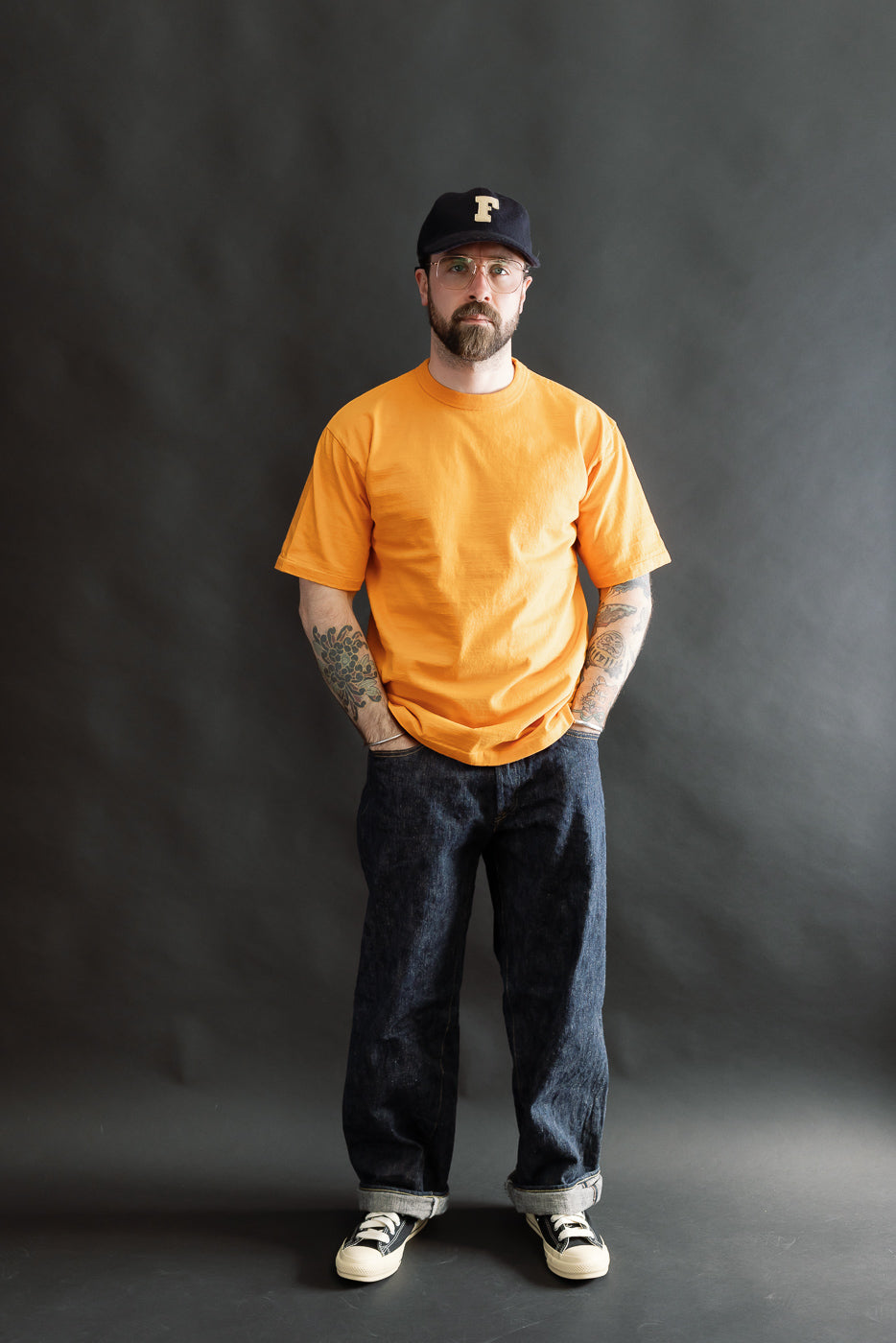 A man models the Fallen Cotton Midweight T-Shirt S/S in Fade Orange, showcasing its casual fit and natural slub texture, paired with dark blue jeans and low-top sneakers.