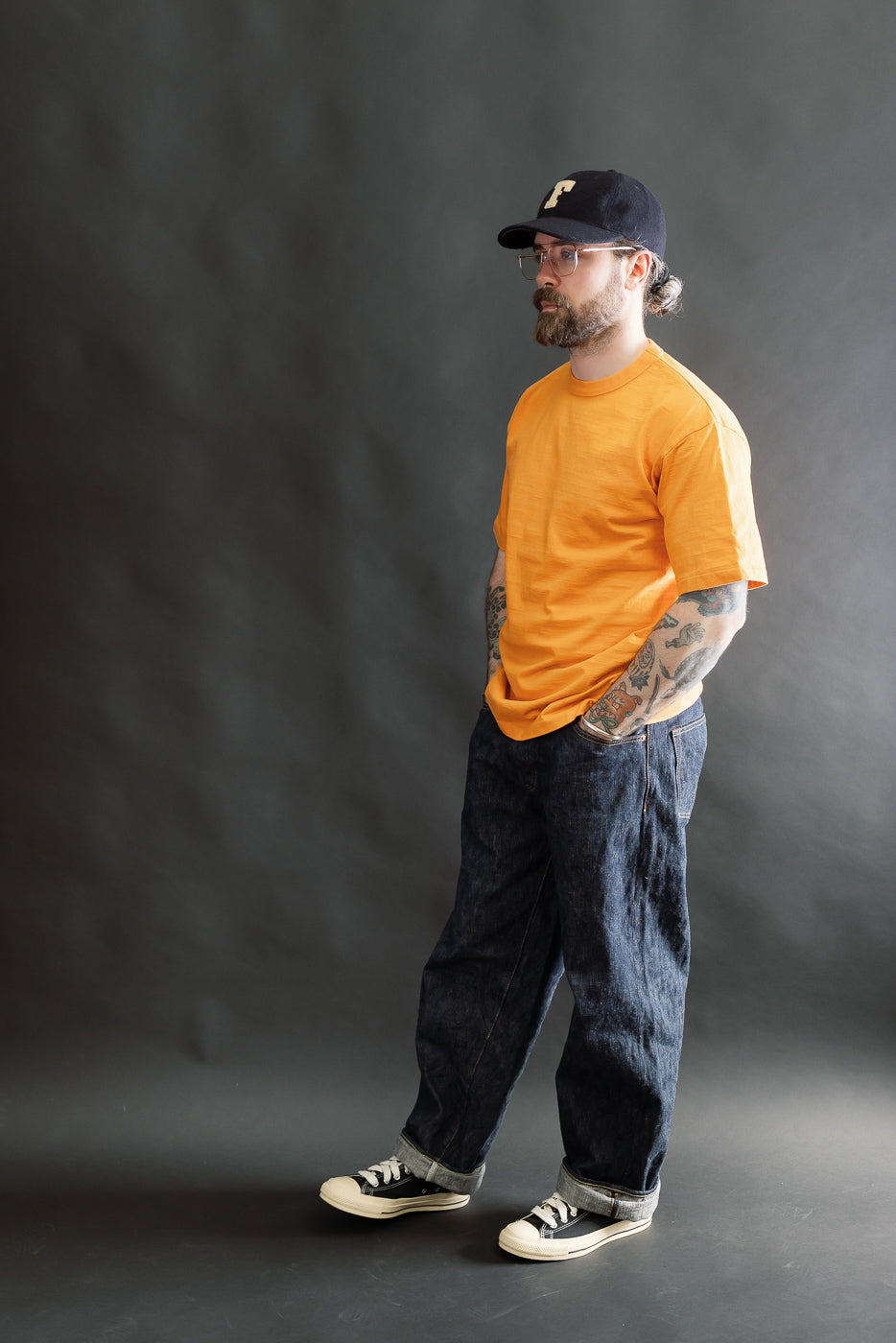 A man models the 7.7oz Fallen Cotton Midweight T-Shirt S/S in Fade Orange, showcasing its relaxed fit and textured recycled cotton slub jersey, paired with dark denim jeans and sneakers.