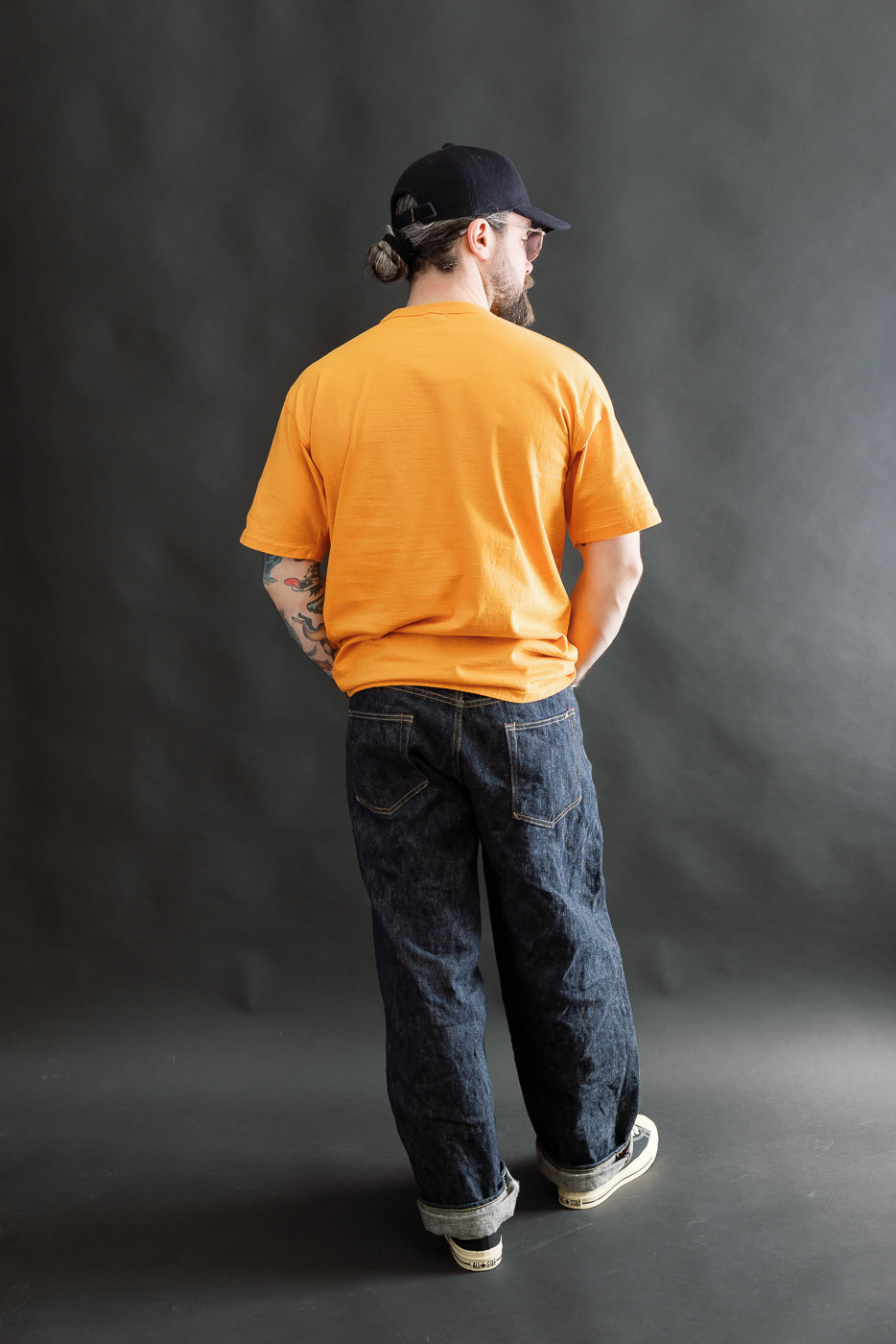 A man models the 7.7oz Fallen Cotton Midweight T-Shirt S/S - Fade Orange, showcasing its relaxed fit and slub texture, paired with dark denim jeans and sneakers, highlighting Dant's heritage-inspired style.