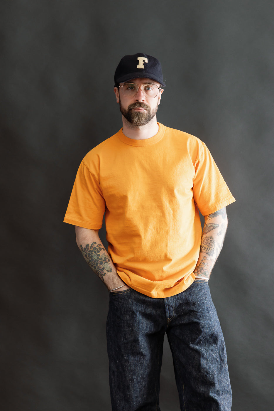 A man models the 7.7oz Fallen Cotton Midweight T-Shirt S/S - Fade Orange, showcasing its relaxed fit and textured recycled cotton slub jersey, styled with jeans and a cap.