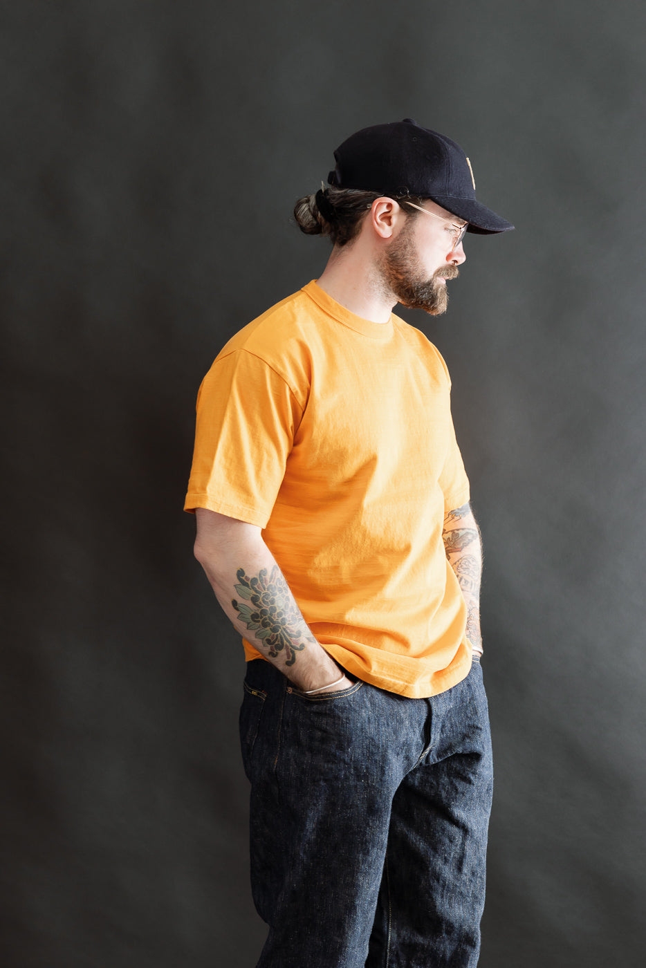 Adult male model wearing the 7.7oz Fallen Cotton Midweight T-Shirt S/S in Fade Orange, styled with dark blue jeans, showcasing recycled cotton's natural slub texture and classic silhouette against a neutral backdrop.