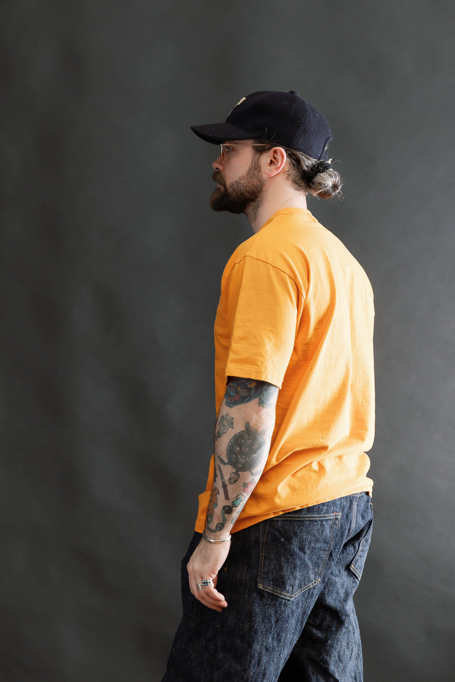 Man wearing 7.7oz Fallen Cotton Midweight T-Shirt S/S - Fade Orange, dark denim jeans, and black baseball cap. The tee features a comfortable, recycled cotton slub jersey texture with a round body and single-stitched hem.